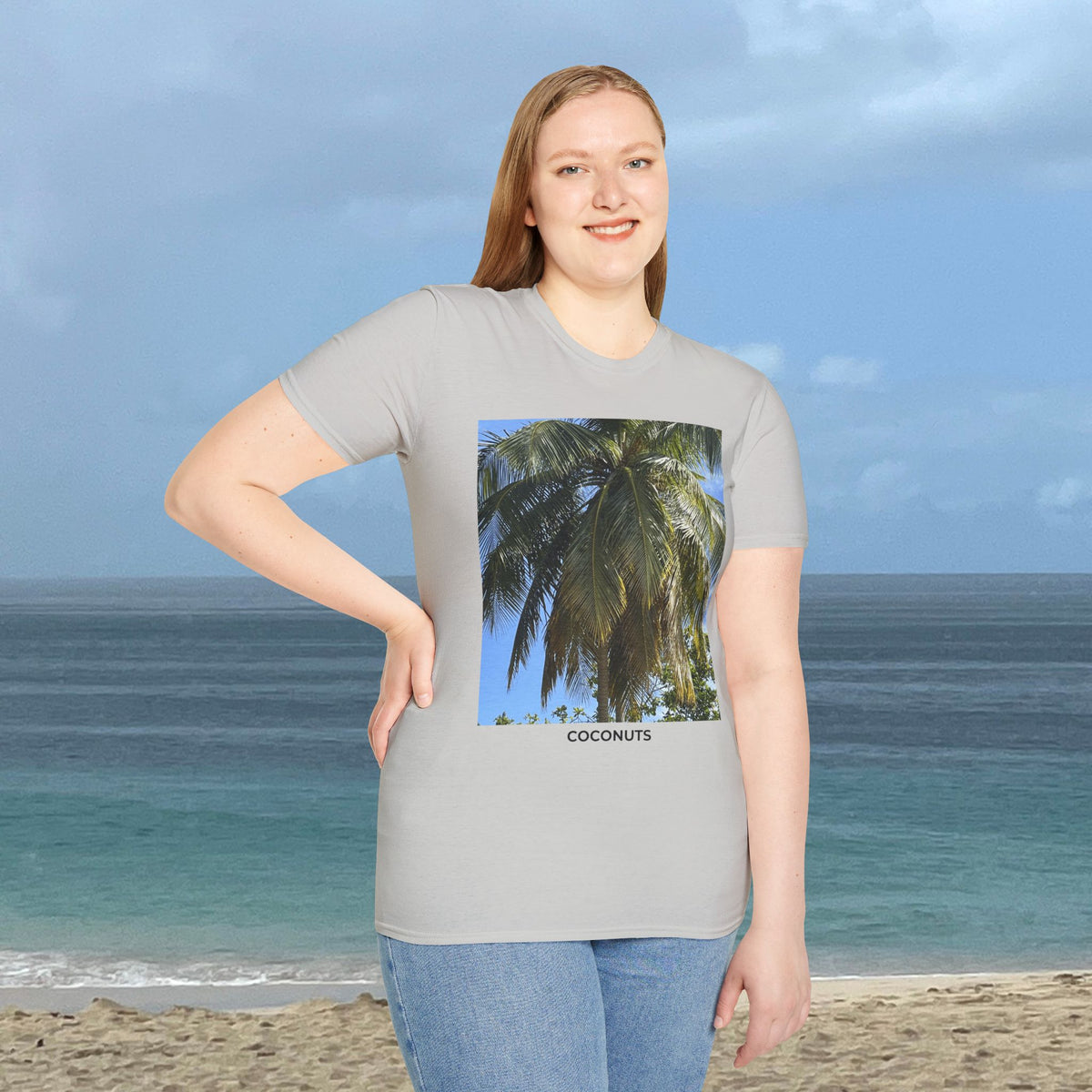 Woman standing by the ocean wearing a light gray Coconut Tree T-shirt with a coconut palm photograph and “COCONUTS” printed on the front.