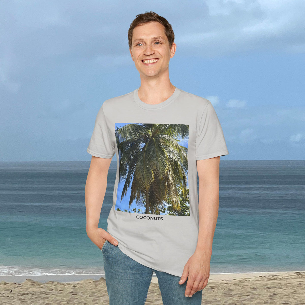 Man standing by the ocean wearing a light gray Coconut Tree T-shirt with a palm photograph and “COCONUTS” printed on the front.