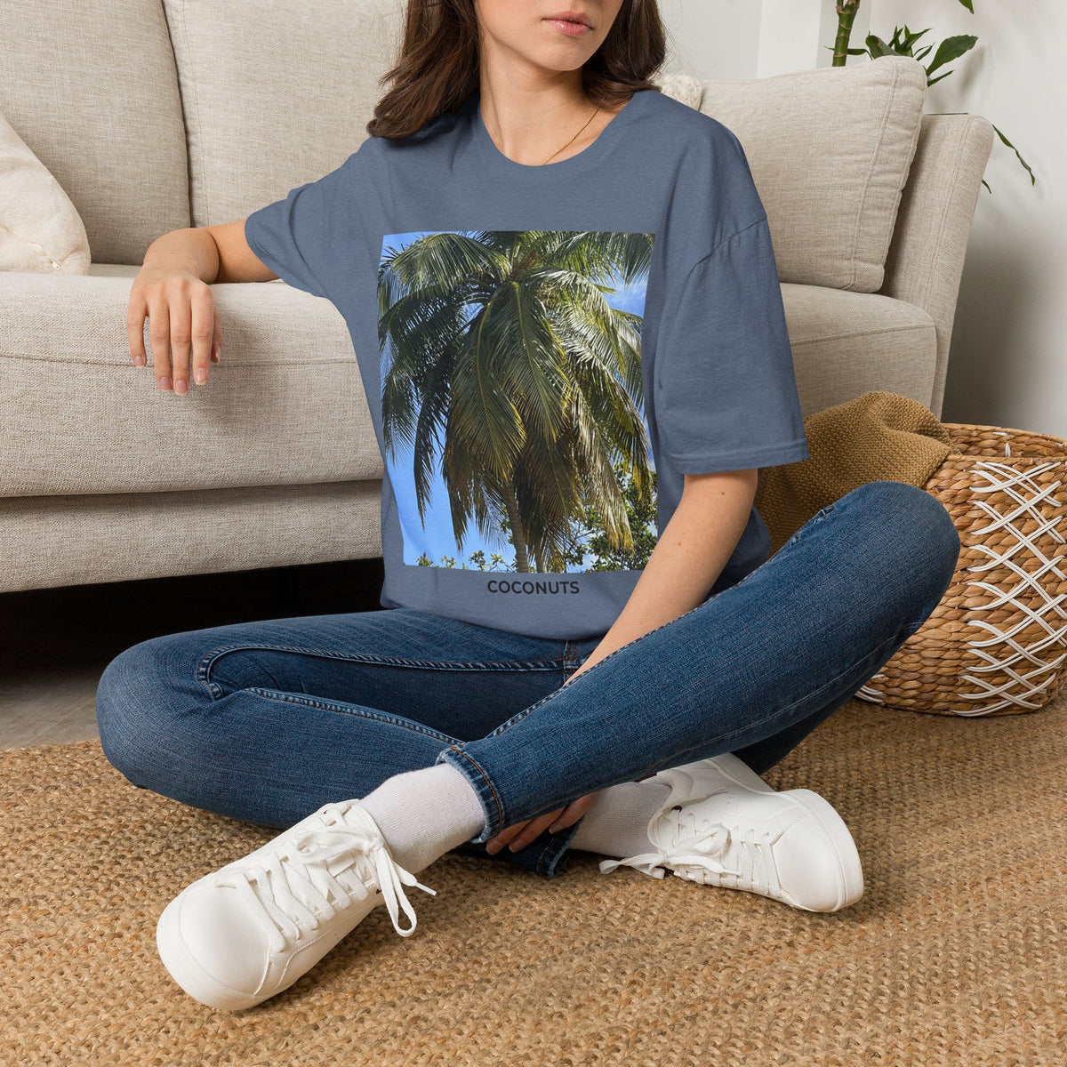 Woman seated indoors wearing a slate blue Coconut Tree T-shirt with a coconut palm photograph and “COCONUTS” printed beneath the image.