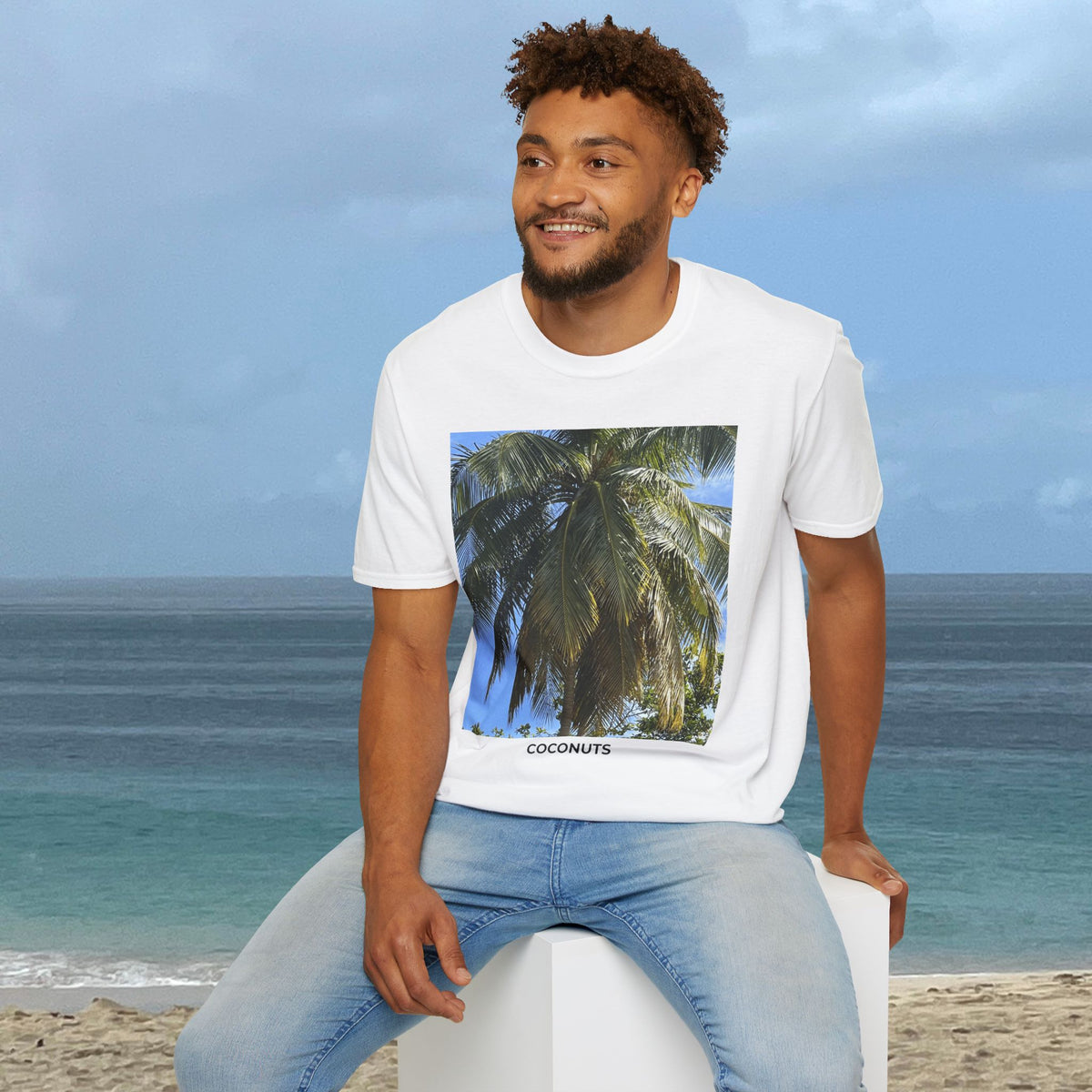 Man seated by the ocean wearing a white Coconut Tree T-shirt with a palm photograph and “COCONUTS” printed on the front