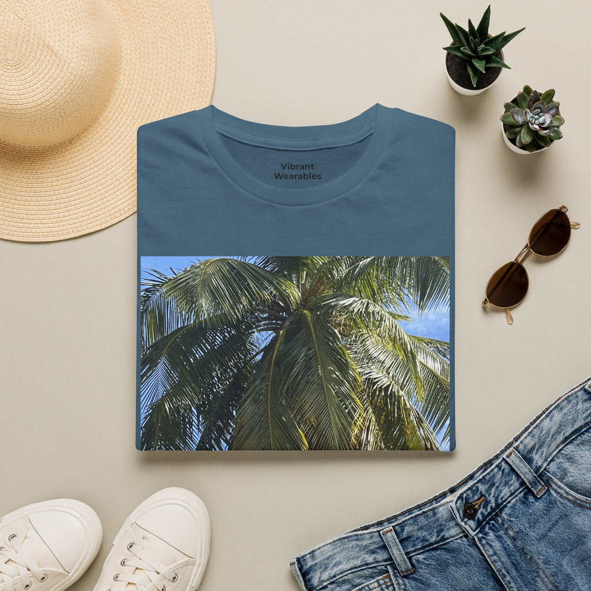Folded indigo blue Coconut Tree T-shirt showing a coconut palm photograph, styled with a straw hat, sunglasses, jeans, and sneakers.
